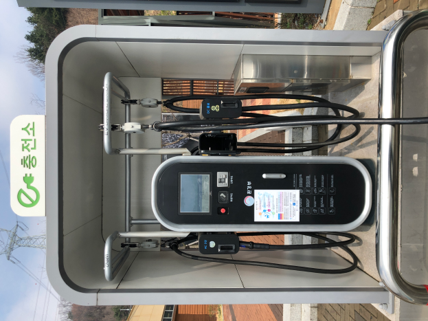 Eine Ladestation für Elektrofahrzeuge mit einem Gerät, Kabeln und einer Texttafel, vor einem Hintergrund aus Gebäuden, Bäumen, Masten, Kabeln und Himmel.