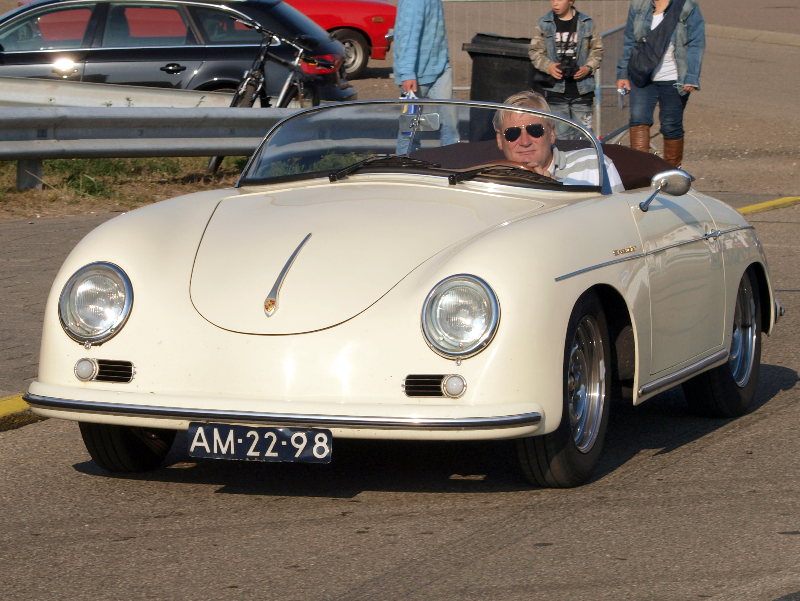 Ein Mann fährt einen weißen Porsche 356 Speedster die Straße entlang, umgeben von Menschen in bunten Kleidern, mit einem Geländer, einem schwarzen Müllcontainer und weiteren Autos und einem Gebäude im Hintergrund.