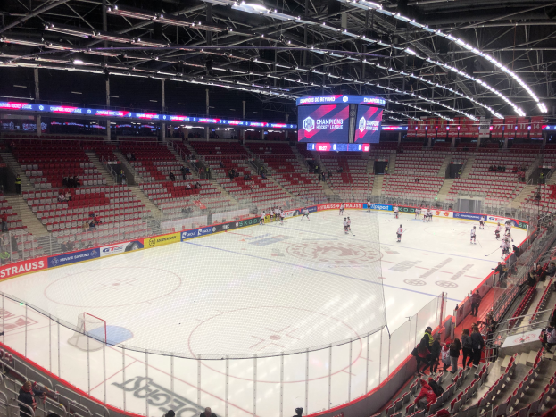 Ein Hockey-Spiel in einer großen Arena mit Zuschauern auf Sitzen und Stehplätzen, einem Zaun um die Spielfläche, einem Bildschirm mit der Aufschrift "St. Louis Blues vs Detroit Red Wings", Metallrahmen und einer Decke mit Leuchten.