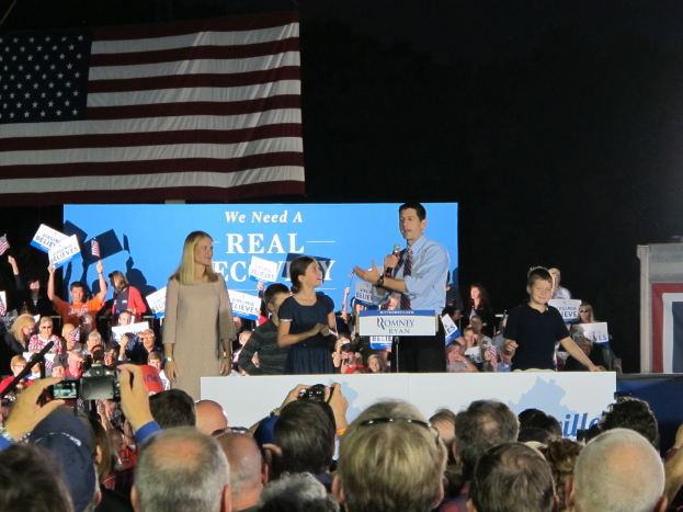 Eine große Menge von Menschen, einige machen Fotos und andere halten Papiere und Fahnen, mit einem Mann in der Mitte, der in ein Mikrofon spricht, und einer Fahne auf der linken Seite.