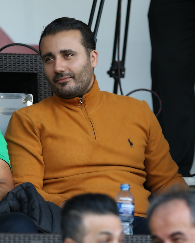Ein Mann in einer Jacke sitzt auf der Tribüne bei einem Tennisspiel, umgeben von Menschen, mit einer Flasche vor ihm und Tribünen und einer Wand im Hintergrund.