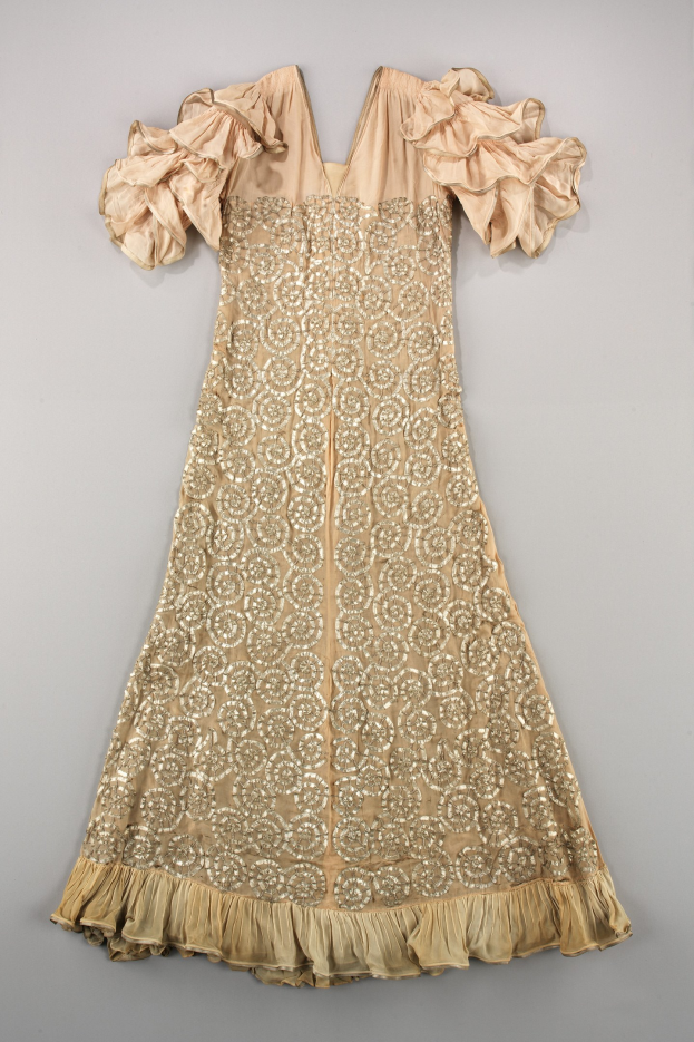 Ein cremefarbenes Kleid mit goldener Stickerei und Rüschen, das auf einer weißen Oberfläche präsentiert wird und feine Designs und Muster aufweist.