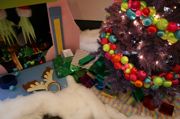 Drinnen steht rechts ein geschmückter Weihnachtsbaum mit Lichterkette und Schmuck, in der Mitte Geschenkboxen und Dekorationen, links oben eine Hütte mit Dekorationen und unten Wolle und Watte.