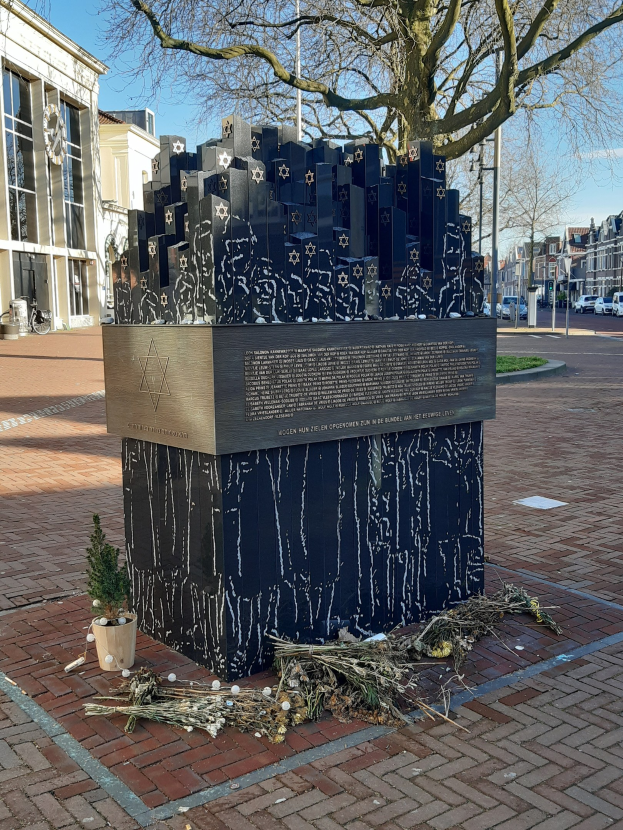 Denkmal in einem Stadtplatz, das an Holocaust-Opfer erinnert, mit einem Baum davor, einem Topfpflanze auf dem Boden, verstreuten trockenen Blättern und Gebäuden, Fahrzeugen, Masten und Bäumen im Hintergrund.