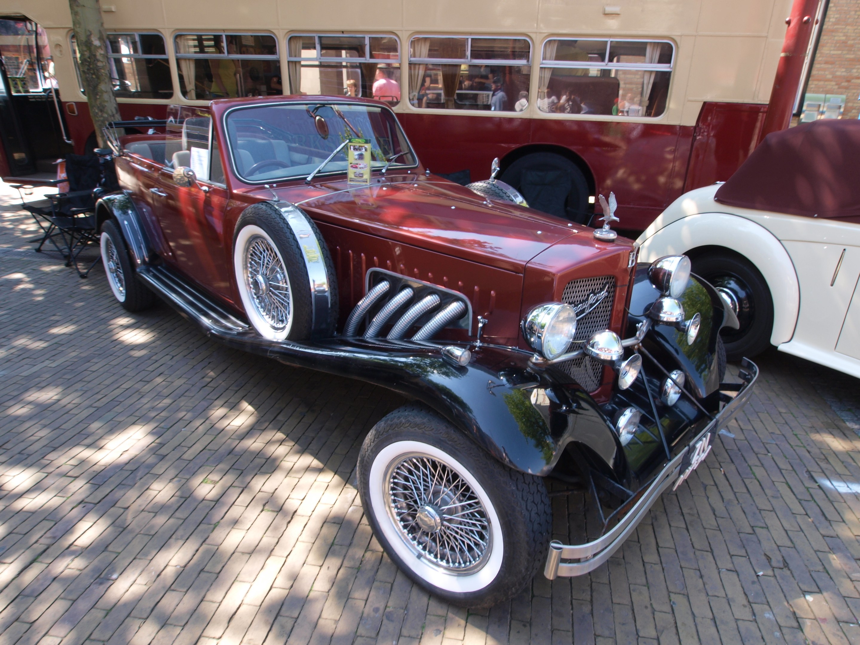 Ein Oldtimer neben einem Doppeldeckerbus auf Kopfsteinpflasterstraße, mit Stühlen und einem Baum im Vordergrund, einem Gebäude mit Fenstern im Hintergrund und Menschen im Bus zu sehen.