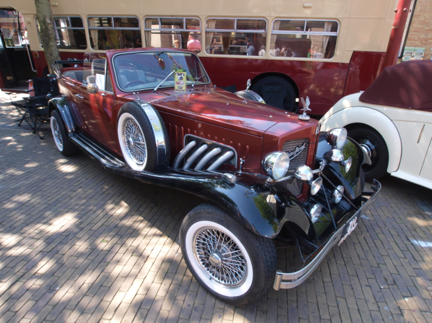 Ein Oldtimer neben einem Doppeldeckerbus auf Kopfsteinpflasterstraße, mit Stühlen und einem Baum im Vordergrund, einem Gebäude mit Fenstern im Hintergrund und Menschen im Bus zu sehen.