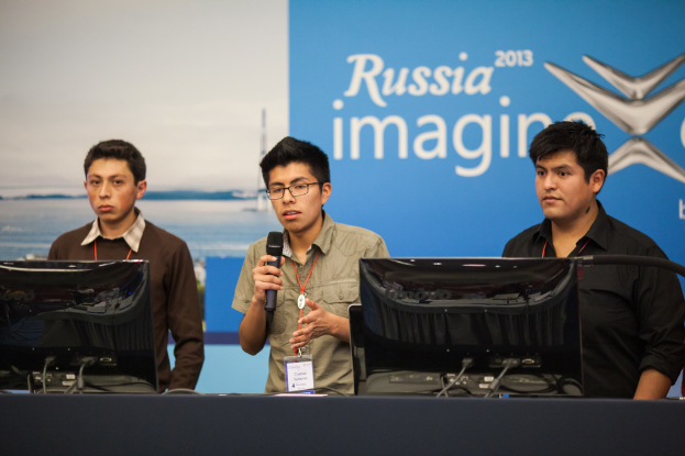 Drei Jungs stehen neben einem Schreibtisch mit zwei Computersystemen, einer spricht in ein Mikrofon, trägt ID-Karten mit roten Markierungen, vor einer Wand mit einer Werbetafel, auf der "Russia Imagine 2013" steht.