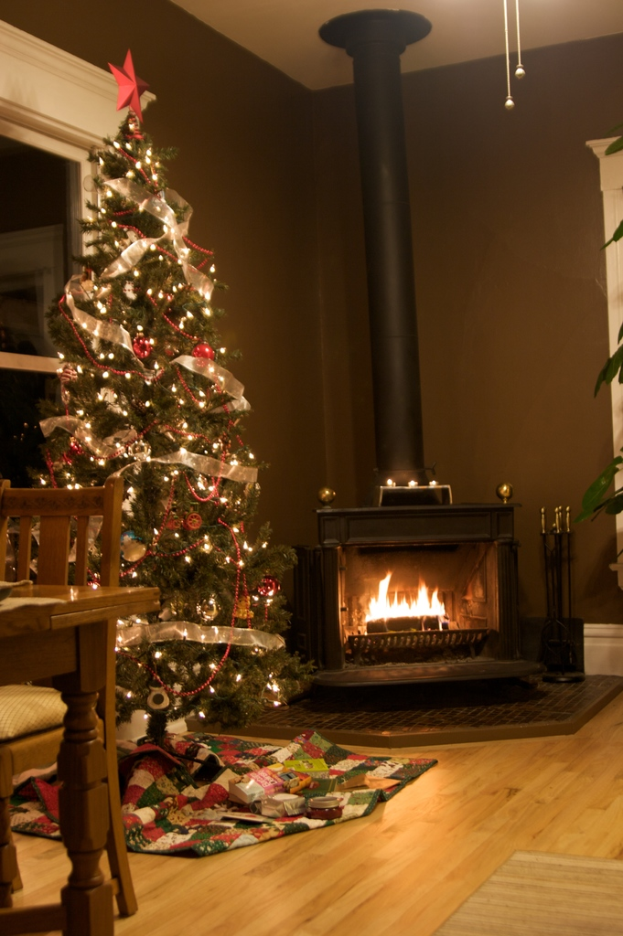 Ein geschmückter Weihnachtsbaum mit Lichtern und Schmuck steht neben einem Tisch und einem Stuhl, mit einem Kamin, einem schwarzen Pfahl, einer Zimmerpflanze, einem Fenster und diversen Gegenständen im Hintergrund.