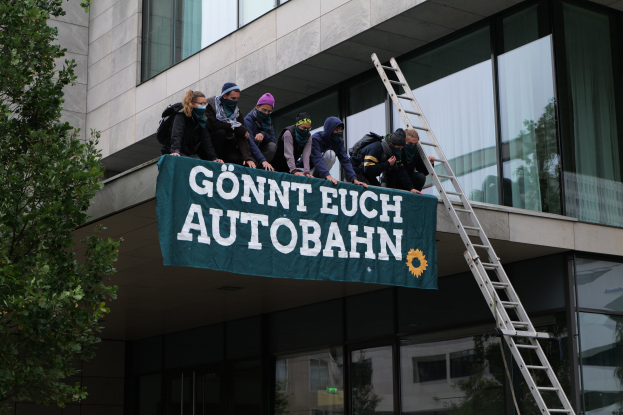 Eine Gruppe von Menschen auf einem Gebäude, die eine Fahne halten, mit einem Baum links und Glasfenstern im Hintergrund, die scheinbar im Support der deutschen Regierung protestieren.