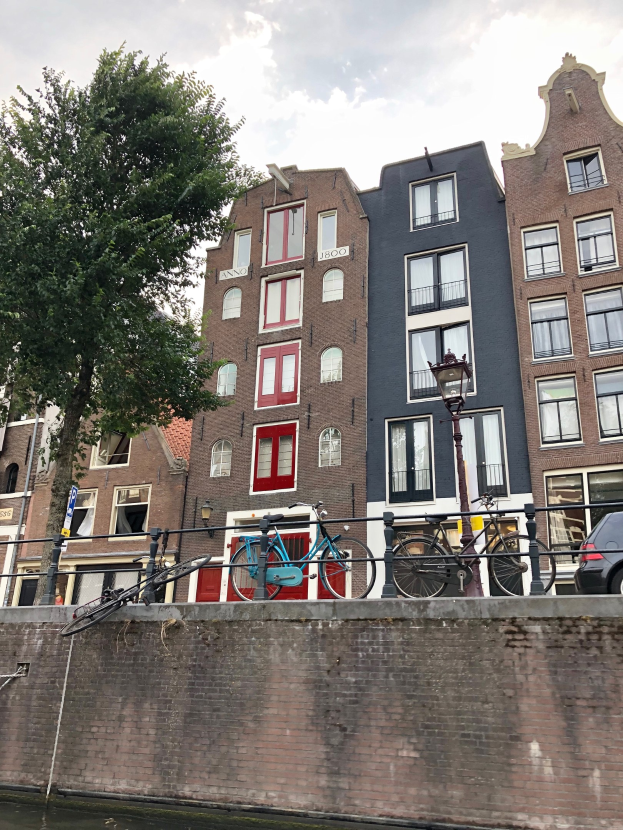 Fahrräder, die an einem Kanal in Amsterdam geparkt sind, mit vorbeifahrenden Fahrzeugen, Gebäuden im Hintergrund und einer bewölkten Landschaft.