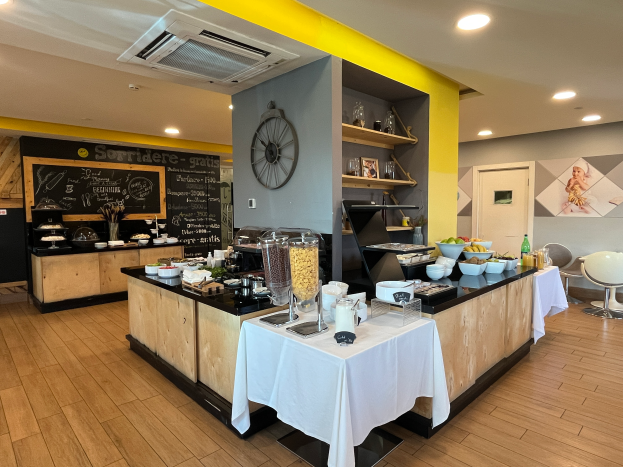 Ein Restaurant-Innenraum mit Tischen und Stühlen, Speisen, einer Tafel mit Text, einer Wanduhr, Deckenleuchten, einer Klimaanlage und einer Tür im Hintergrund, im Hotel de Ville in Paris gelegen.