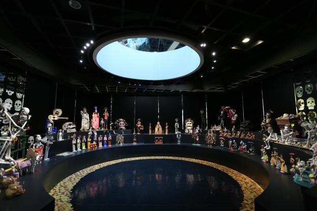 Ein großer Raum voller verschiedener Spielzeuge, darunter Skelette, die auf dem Boden angeordnet sind, mit Totenschädeln an den Wänden und Lichtern an der Decke, die eine festliche Atmosphäre für das Tag der Toten-Fest schaffen.