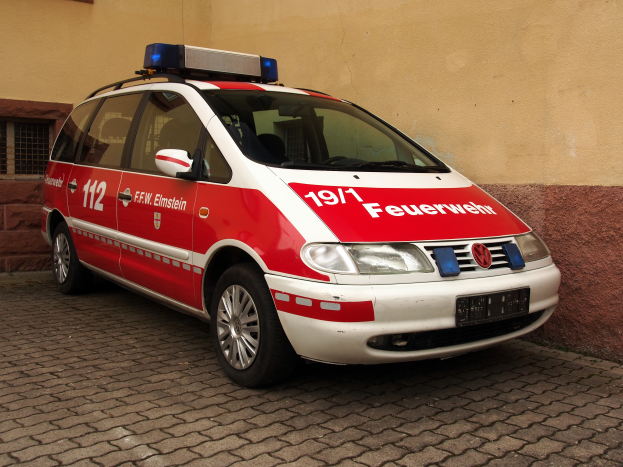 Ein roter und weißer Minivan, der als Krankenwagen mit Text und Zahlen gekennzeichnet ist, steht vor einem Gebäude mit Fenstern.