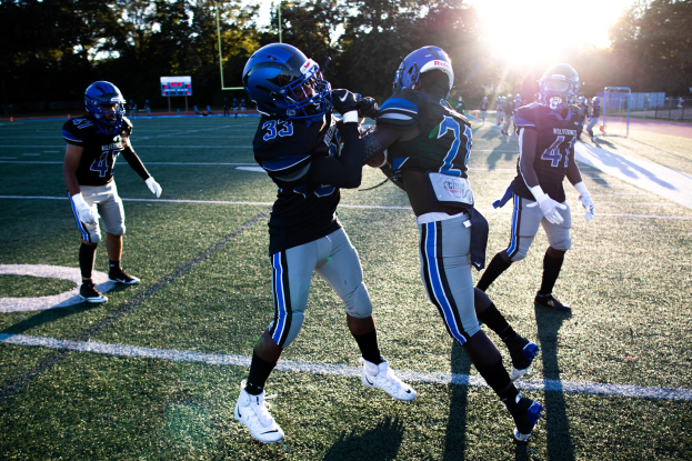 Gruppe junger Männer in Helmen und Schuhen beim Fußballspielen auf einem Feld mit Bäumen und einem klaren blauen Himmel.