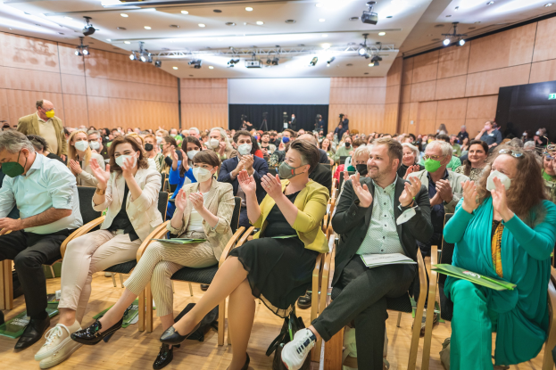 Eine Gruppe von Menschen sitzt in Stühlen vor einem Publikum, einige tragen Masken, mit Taschen auf dem Boden; ein wandmontierter Bildschirm und Deckenleuchten deuten auf eine Coronavirus-Konferenz hin.