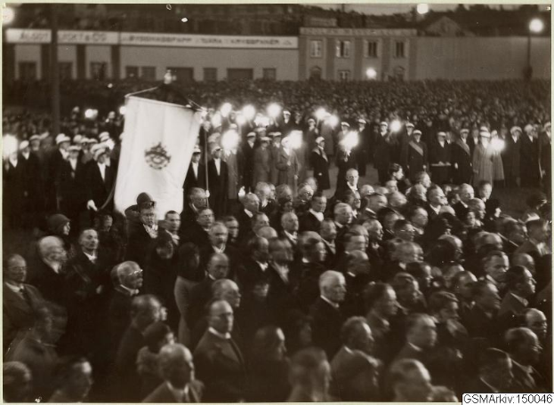 Schwarzes und weißes Foto einer großen Menschenmenge vor einem Gebäude, mit einer Person, die eine Fahne hält, sichtbaren Masten und Lichtern im Hintergrund und Text unten, der "Gustav Klimts Rede an der Berliner Mauer" lautet.
