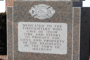Denkmstein mit einer Plakette für die Feuerwehrleute in Millbury, vor einem Backsteingebäude mit umliegender Vegetation.