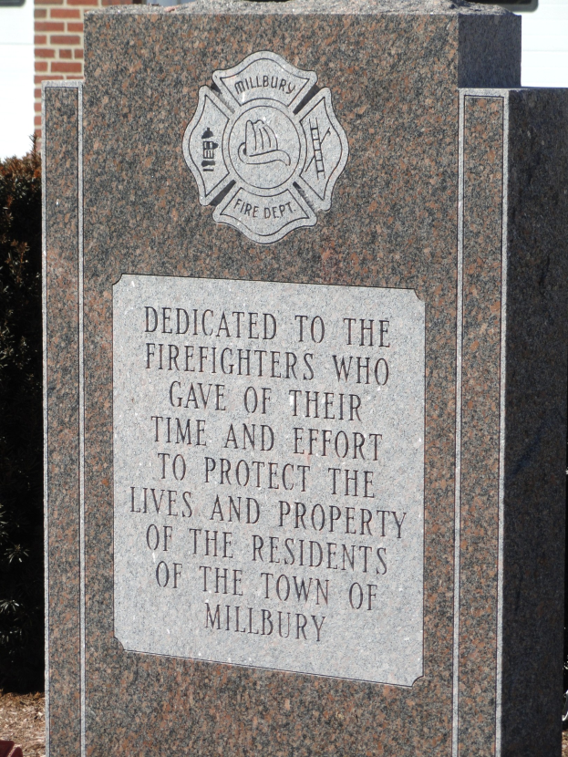Denkmstein mit einer Plakette für die Feuerwehrleute in Millbury, vor einem Backsteingebäude mit umliegender Vegetation.