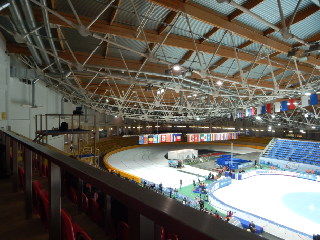 Große Indoor-Eisbahn mit zahlreichen Menschen, die Schlittschuh laufen, umgeben von Sitzplätzen, Geländern, Brettern, Leuchten und strukturellen Elementen, mit einer Wand im Hintergrund.