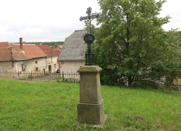 Ein hölzernes Kreuz steht als Holocaust-Mahnmal in einem von einem Metallzaun umgebenen Grasfeld, mit Bäumen, Häusern und einem bewölkten Himmel im Hintergrund.