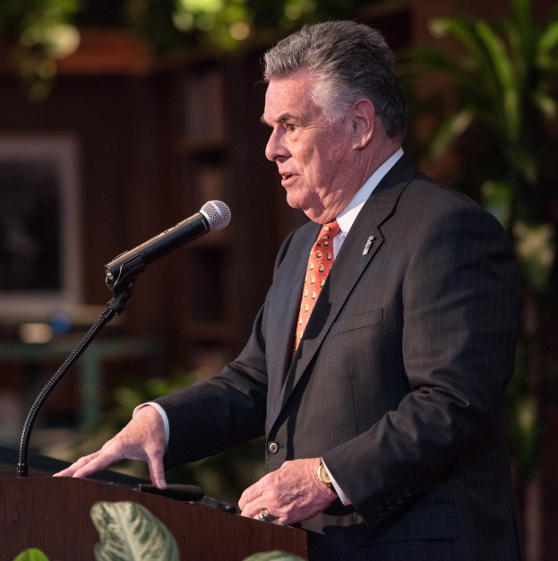 Ein Mann im Anzug und Krawatte steht an einem Podium mit einem Mikrofon, umgeben von Blättern mit einem unscharfen Hintergrund.
