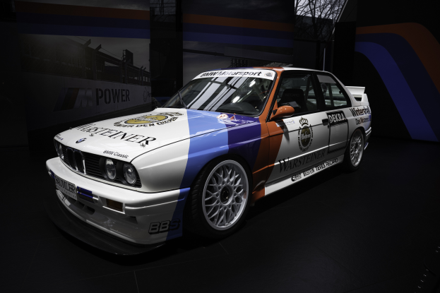 Ein BMW E30 M3 Motorsportwagen ist auf dem Boden mit Text darauf zu sehen, umgeben von Bannern mit Text und Bildern im Hintergrund, mit einem Rad in der Mitte des Bildes.
