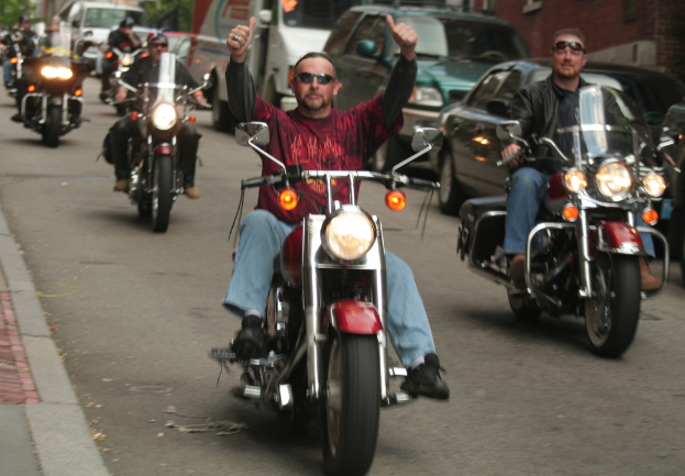 Eine Gruppe von Menschen auf Fahrrädern auf einer Straße, wobei die Person in der Mitte ein rotes Hemd trägt und ohne Hände fährt, und eine andere Person rechts mit einer schwarzen Jacke und einem Lachen. Im Hintergrund sind zwei Autos und ein Lieferwagen zu sehen.
