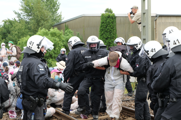 Eine Gruppe von Polizisten in schwarzen Uniformen und weißen Helmen steht um einen Mann in einem weißen Kleid auf einem Bahngleis, mit Bäumen, einem Gebäude und einem klaren blauen Himmel im Hintergrund.