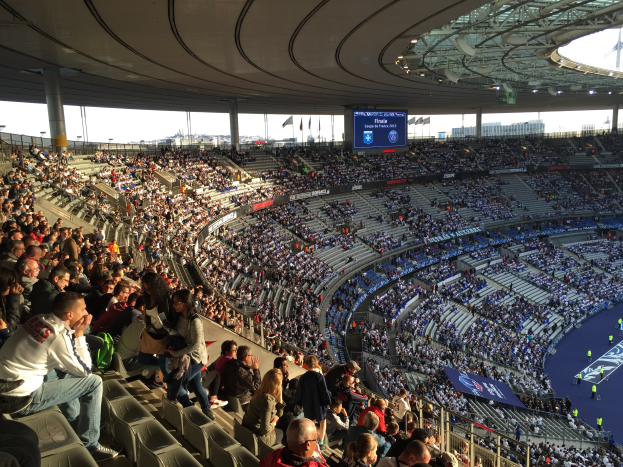 Große Menschenmenge in einem Stadion bei einem Fußballspiel, mit einer Bühne rechts, Fahnen, Stangen, einem Bildschirm und der Allianz Arena in München, Deutschland.