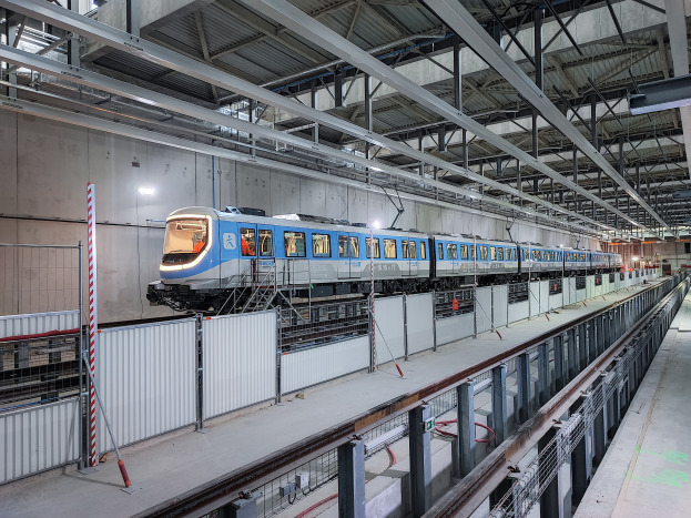 Ein blau-weißer Zug fährt durch ein großes Gebäude mit einer Plattform auf der rechten Seite, einer Wand im Hintergrund und einer Decke mit Stangen und Lampen oben, was darauf hinweist, dass es Teil eines neuen Bahnsystems in Deutschland ist.