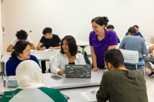 Eine Gruppe von Menschen in einem Klassenzimmer, die um einen Tisch mit Laptops, Büchern und anderen Gegenständen sitzen und über Klimawandel diskutieren.