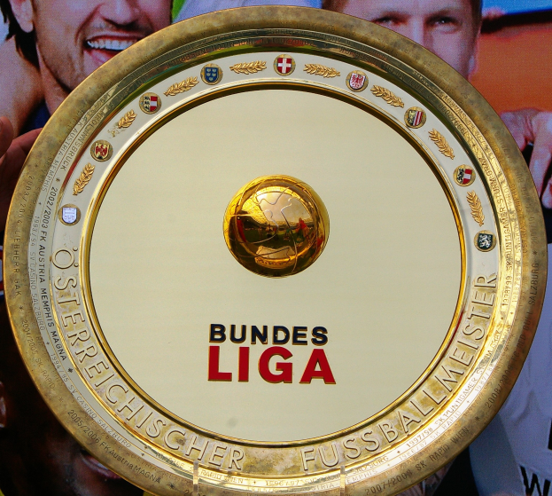 Goldener Schild mit "Bundesliga"-Text in Händen eines Mannes, mit einem Banner mit drei Personen im Hintergrund.