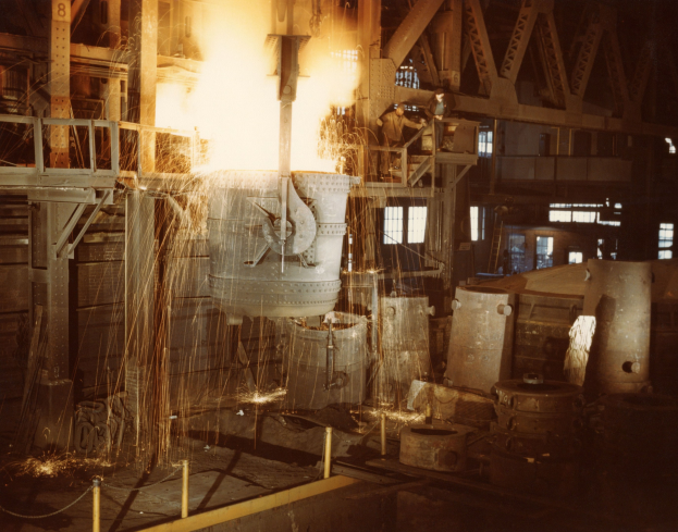 Großes Metallobjekt wird in einen Ofen gegossen, Menschen stehen drumherum, Feuer erhellt die Gegend, Metallobjekte, Pfähle und Fenster im Hintergrund.