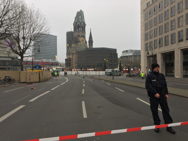 Polizeibeamter in schwarzer Uniform und Mütze auf einer Berliner Straße mit Verkehrskegeln, Fahrrädern, Laternen, Bäumen und Gebäuden, mit einem Uhrenturm und blauem Himmel im Hintergrund.
