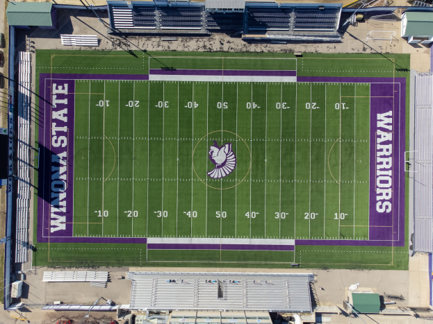 Eine Luftaufnahme eines Footballfelds mit violetten und weißen Markierungen, umgeben von Gebäuden, Fahrzeugen und Masten, zeigt das Heimfeld der Minnesota State University Footballmannschaft.