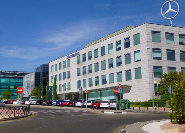 Die Mercedes-Benz-Zentrale in Frankfurt, Deutschland, mit Fahrzeugen auf der Straße, Schildern, Laternenpfählen, Bäumen, Pflanzen und einem Geländer im Vordergrund, unter einem bewölkten Himmel.