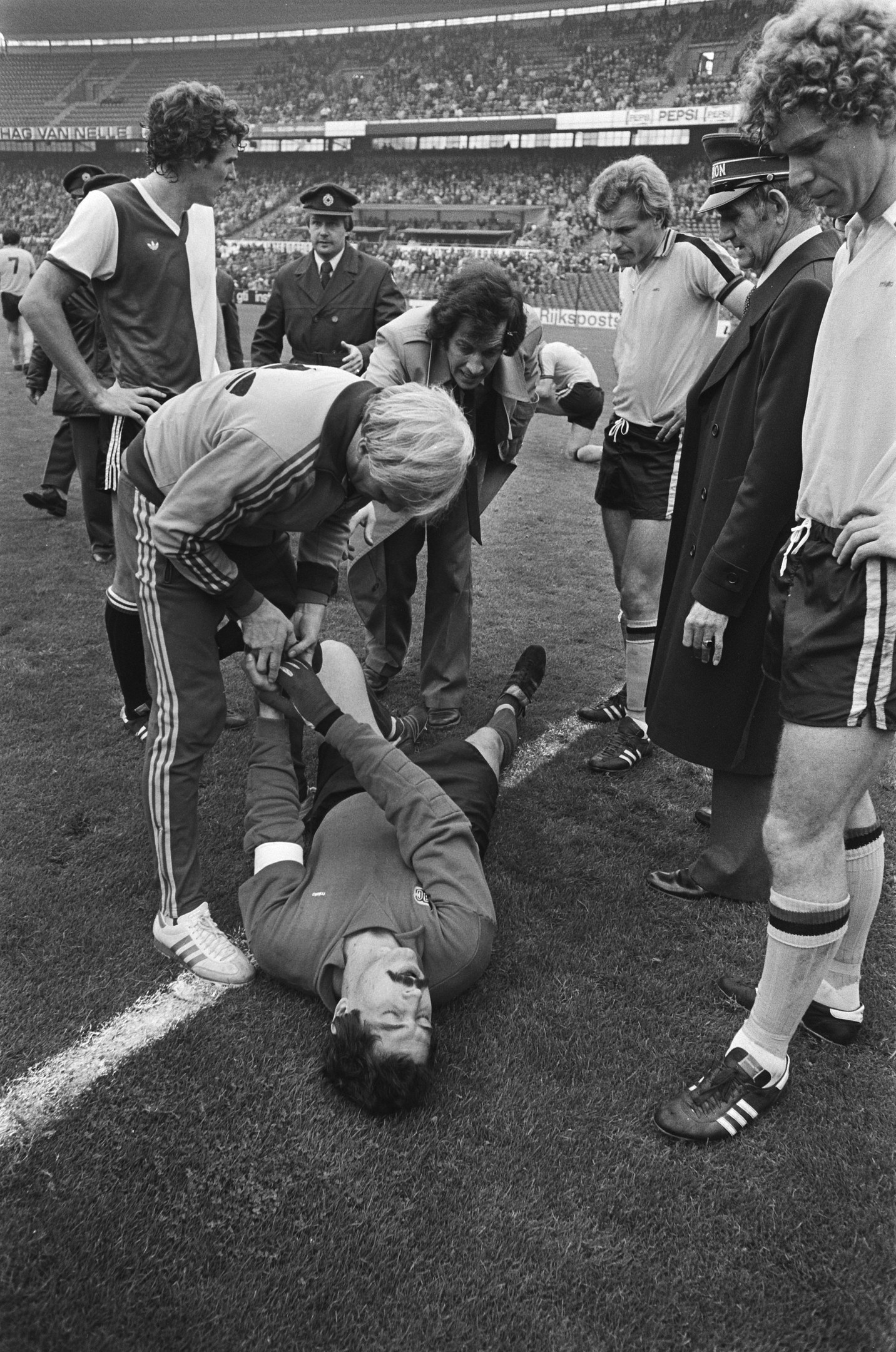 Schwarzes und weißes Bild eines Fußballspielers auf dem Boden umgeben von einer Gruppe von Männern, mit einer Menschenmenge im Hintergrund der Arena.