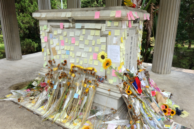 Denkmal mit Post-it-Notizen und Blumen, umgeben von Bäumen, Pflanzen und Gras in einer friedlichen Umgebung.
