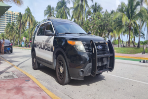 Polizeiauto am Straßenrand mit Menschen, Fahrrädern, Gras, Bäumen, Gebäuden und einem klaren blauen Himmel im Hintergrund.