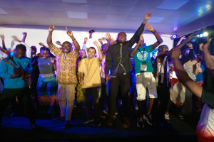 Eine Gruppe von Menschen auf einer Bühne mit Mikrofonen, Musikinstrumenten und einem Vorhang im Hintergrund, die Hände klappend in Feierlaune