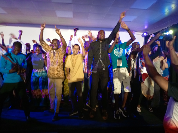 Eine Gruppe von Menschen auf einer Bühne mit Mikrofonen, Musikinstrumenten und einem Vorhang im Hintergrund, die Hände klappend in Feierlaune