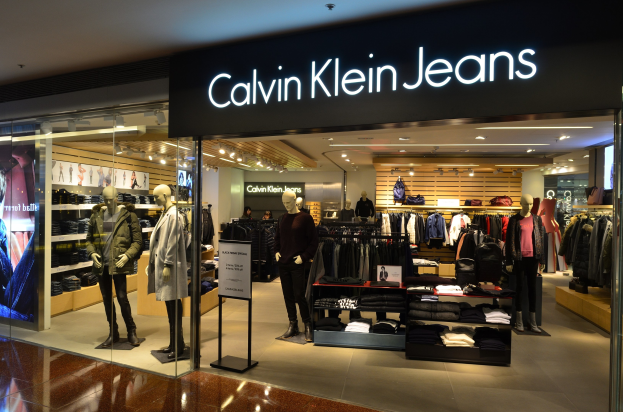 Inneneinrichtung eines Calvin Klein Jeans-Geschäfts in einem Einkaufszentrum, das Schaufensterpuppen in Kleidungsauslagen, hängende Kleidung, eine Werbetafel, einen digitalen Bildschirm und eine Deckenbeleuchtung zeigt.