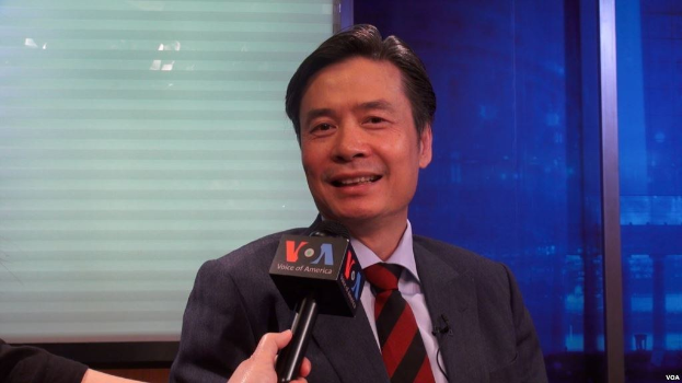 Ein Mann im Anzug, der ein Mikrofon hält, vor einem Fenster und einer Wand.