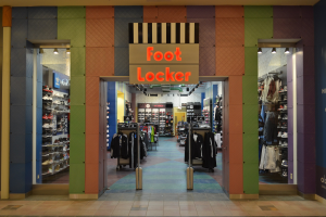 Innenansicht eines Foot Locker Schuhgeschäfts mit Kleidung an Bügeln, Waren auf Gestellen und Deckenbeleuchtung, die den Ladenboden zeigt.
