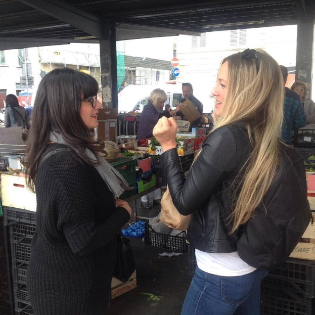 Zwei Frauen stehen und sprechen, halten eine Abdeckung, mit einigen Menschen und Gegenständen im Hintergrund unter einem Dach und Gebäuden weiter hinten.