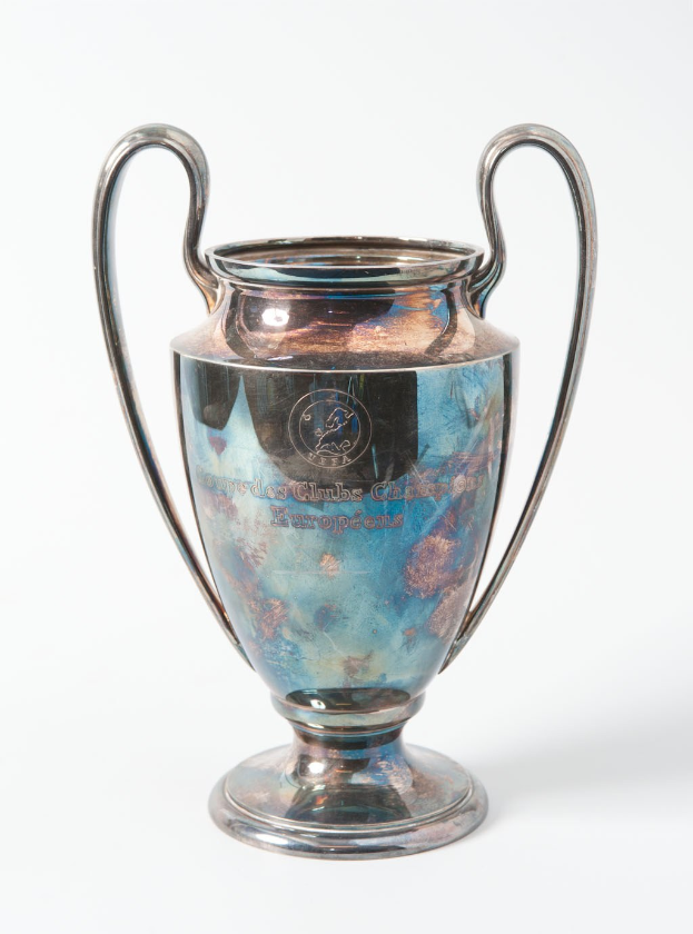 Der UEFA Champions League Pokal, ein stählerner Becher mit zwei Griffen, blau und silbern, mit der Inschrift 'UEFA Champions League', vor einem weißen Hintergrund.