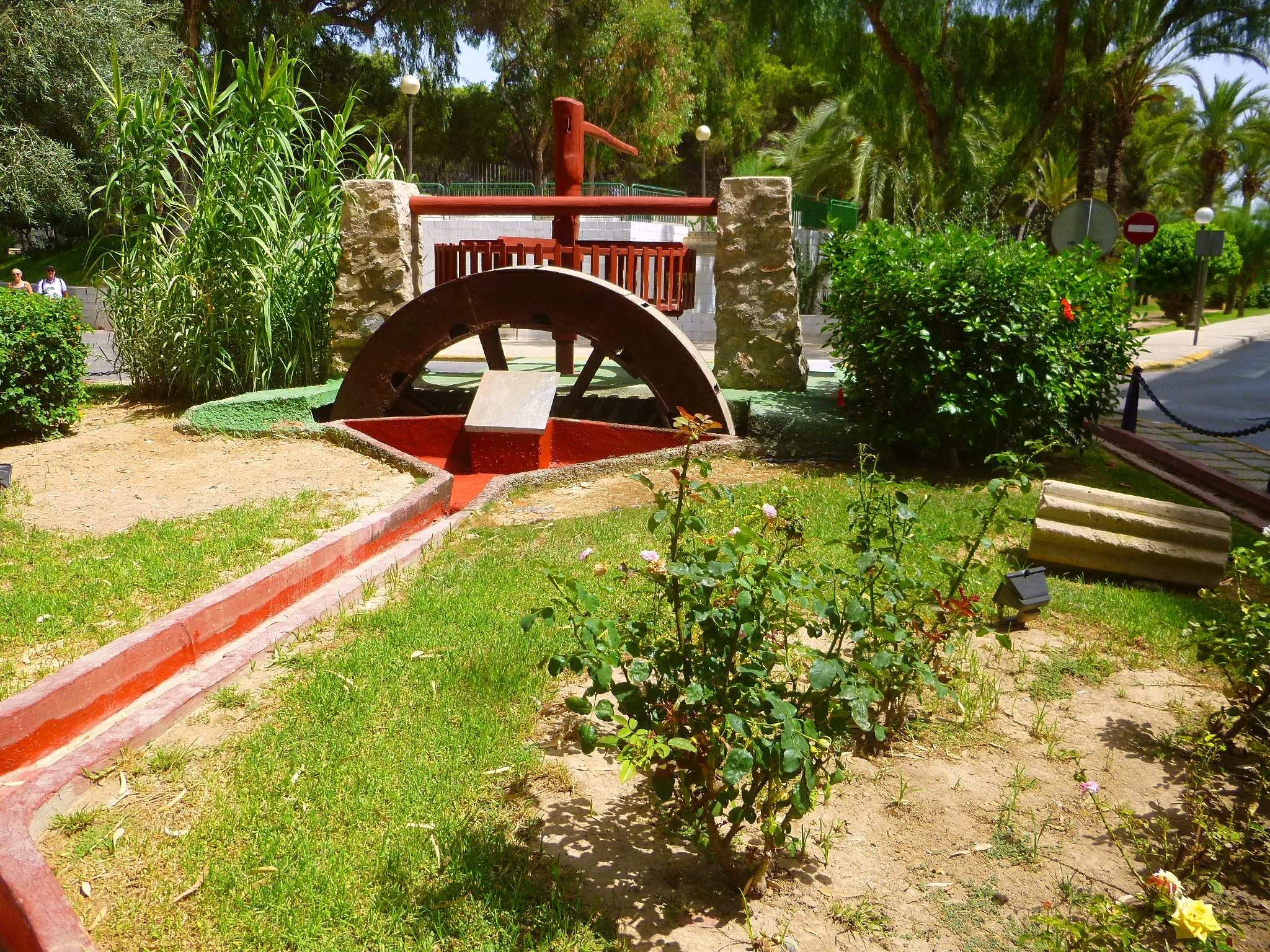 Minigolfanlage in einem Park mit Grünflächen, einer Straße mit Fußgängern, Hinweisschildern und einem klaren Himmel im Hintergrund.