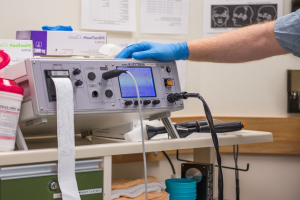 Eine Person in blauen Handschuhen arbeitet an einem medizinischen Gerät auf einem Tisch umgeben von verschiedenen Gegenständen, mit Papieren an der Wand in einer Krankenhausumgebung.