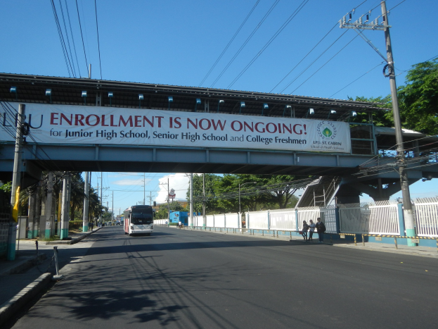 Ein Bus fährt unter einer Brücke mit einem Schild, auf dem "Jetzt Anmelden" steht, Menschen gehen auf einem Fußweg darunter, Strommasten mit Drähten entlang der Straße, Bäume und ein klarer blauer Himmel im Hintergrund.
