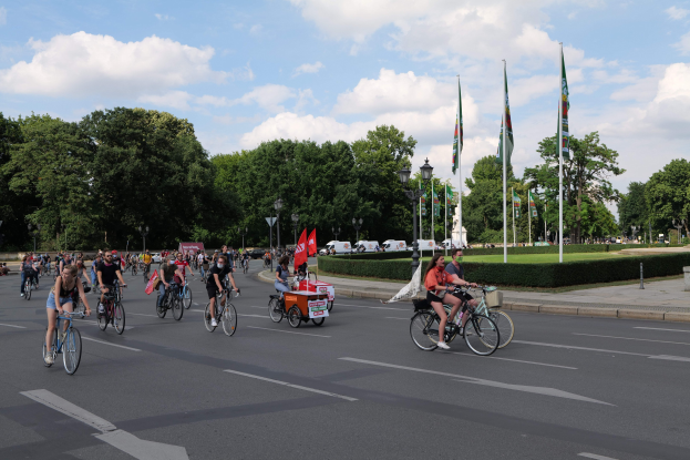 Eine Gruppe von Menschen, die auf Fahrrädern eine von Bäumen gesäumte Straße entlangfahren, während im Hintergrund Laternenpfähle, Fahnenmasten, eine bewölkte Sonne und Fahrzeuge zu sehen sind.